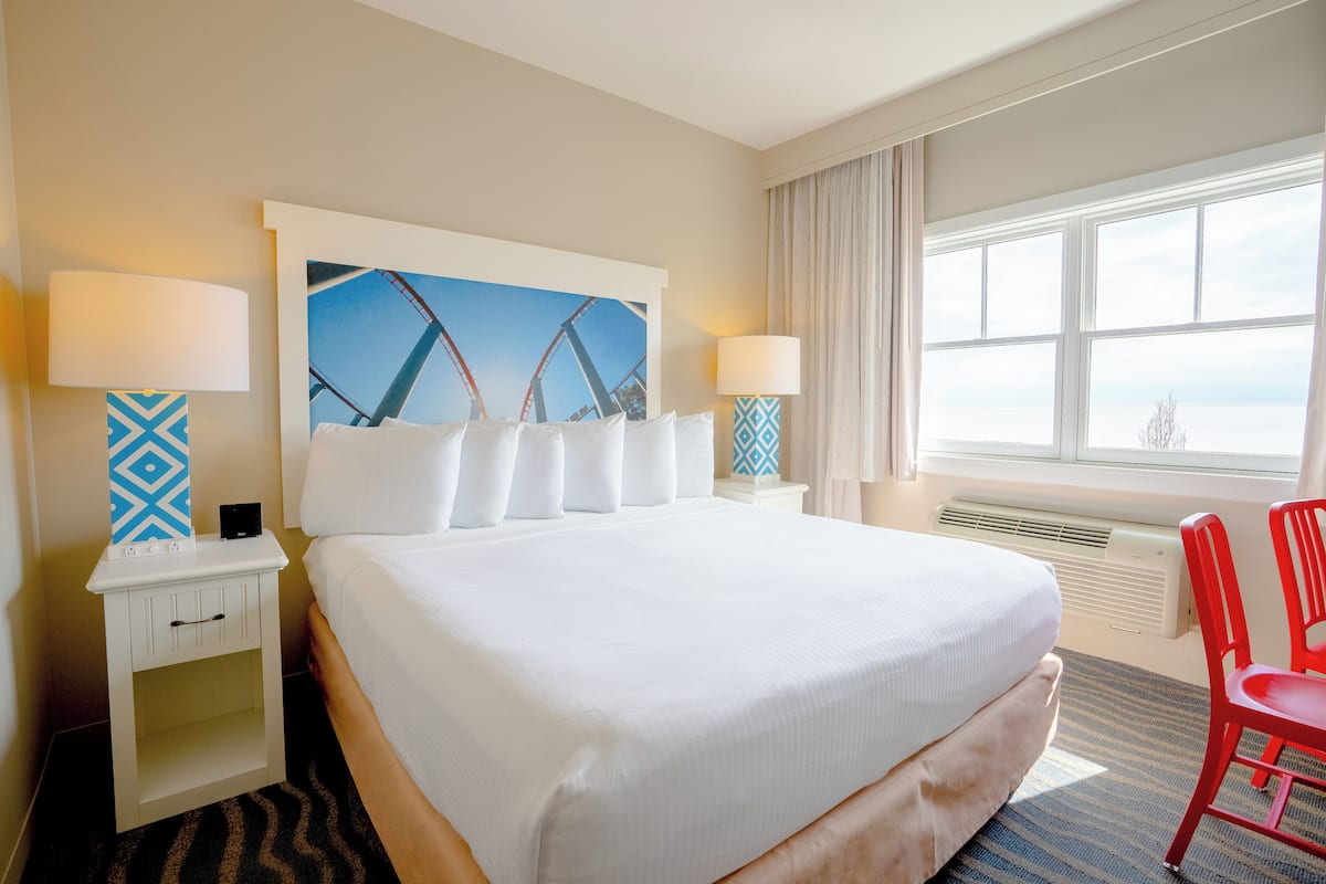 Habitación de hotel con una cama grande, dos mesitas de noche con lámparas, un cuadro enmarcado en la pared, una ventana con cortinas y una silla roja.