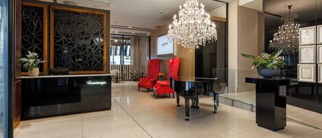 Um hall moderno com balcão de atendimento, cadeiras vermelhas, um piano de cauda e um lustre.
