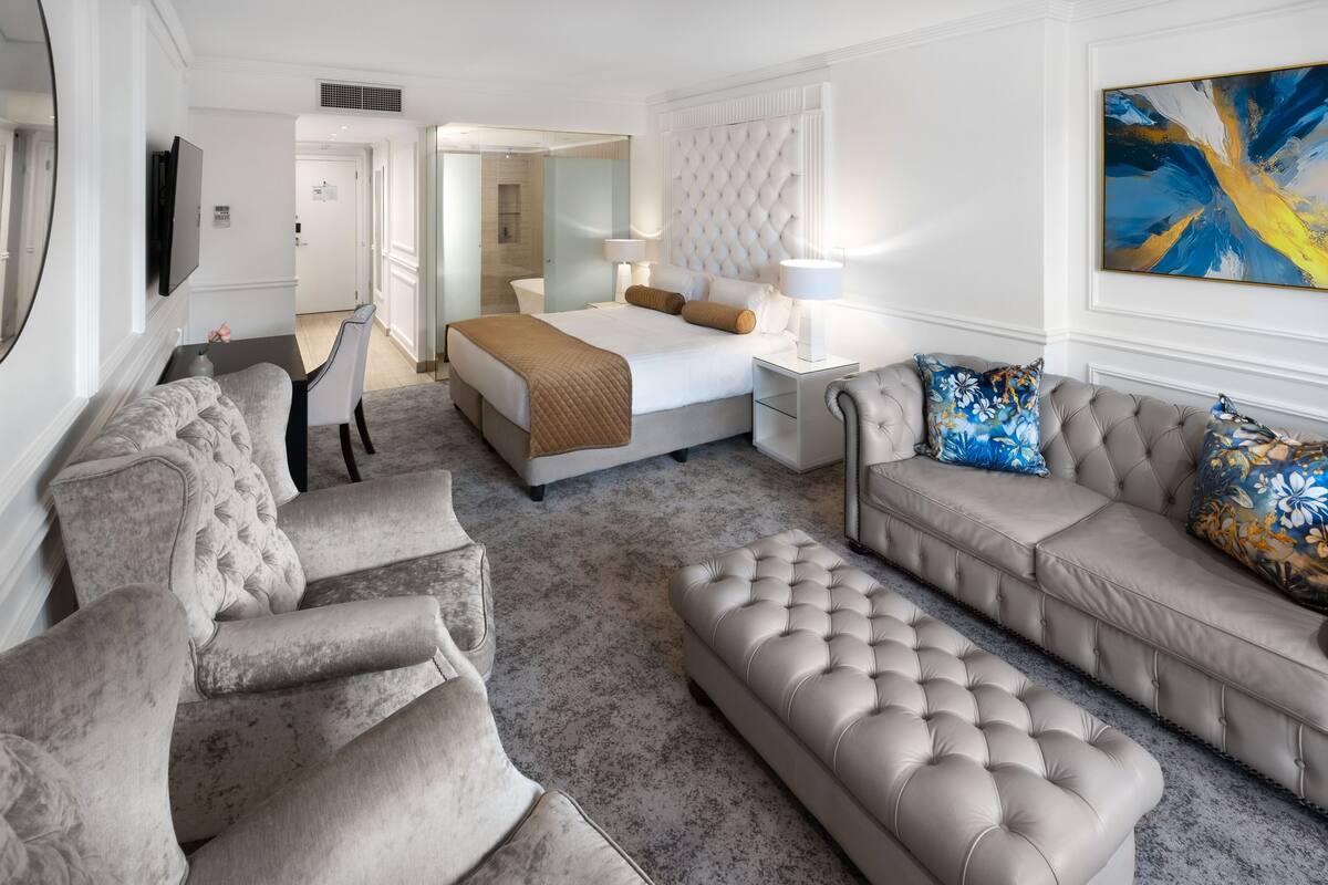 Una habitación de hotel moderna con una cama grande, una zona de estar con sofá y sillón, un escritorio pequeño y un ventanal amplio.