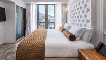 A hotel room with a large bed, a wooden nightstand, a flat-screen TV, and a view of a cityscape through a large window.