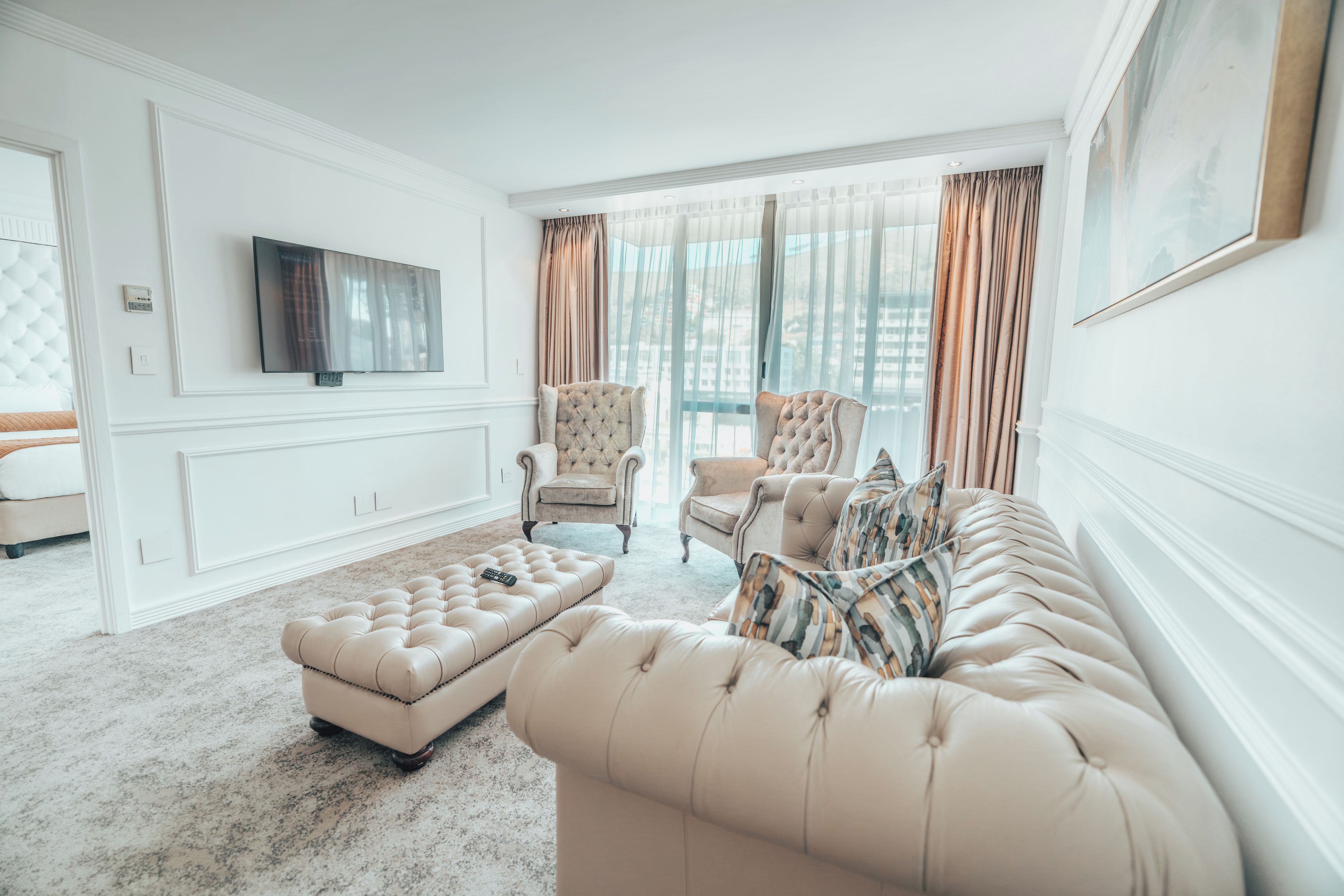 A spacious living room with a large sofa, a tufted ottoman, and two armchairs. A flat-screen TV is mounted on the wall, and the room has large windows with curtains.
