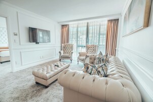 A spacious living room with a large sofa, a tufted ottoman, and two armchairs. A flat-screen TV is mounted on the wall, and the room has large windows with curtains.