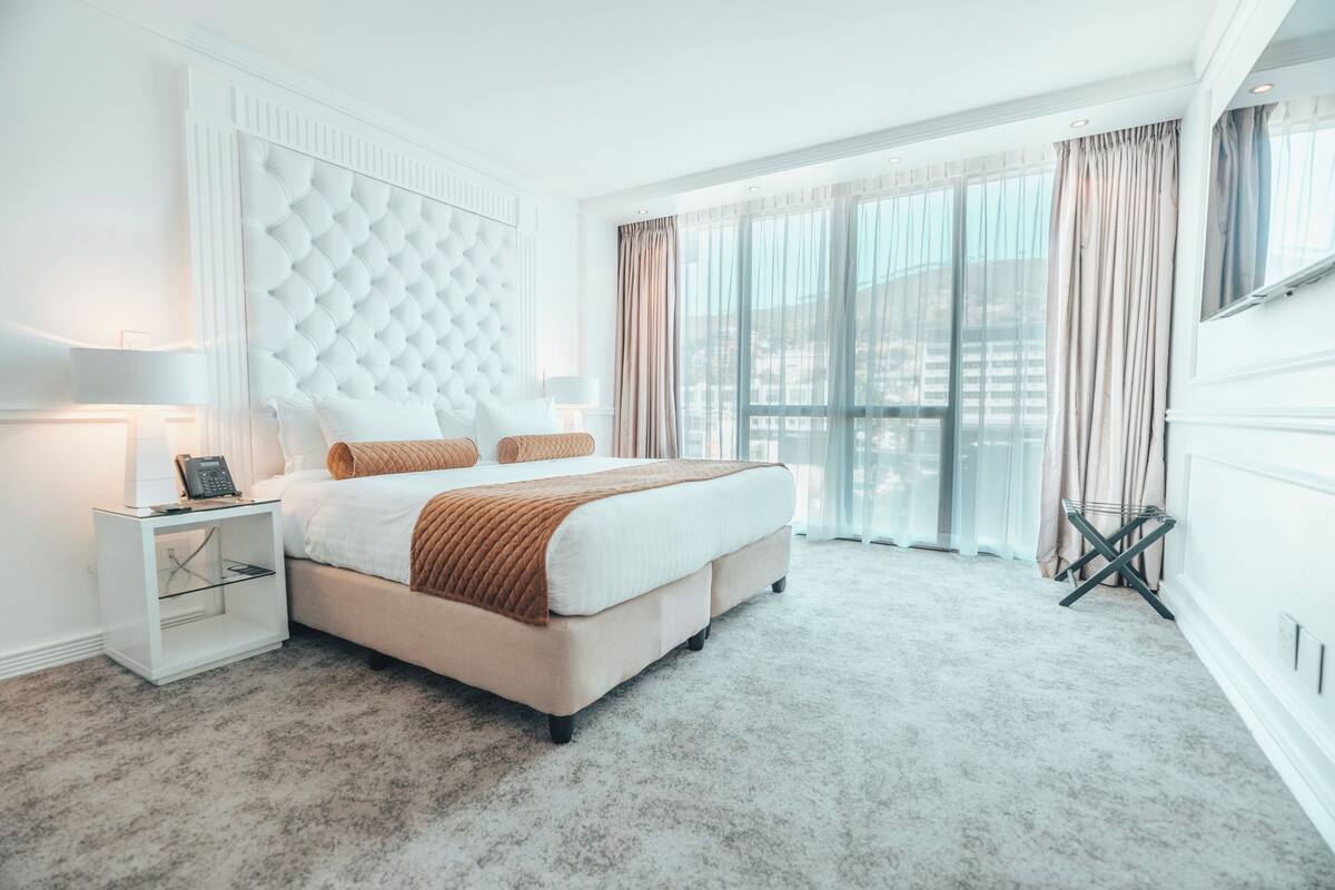 Habitación de hotel moderna con una cama grande, una mesita de noche, una lámpara y vistas de la ciudad a través de amplias ventanas.