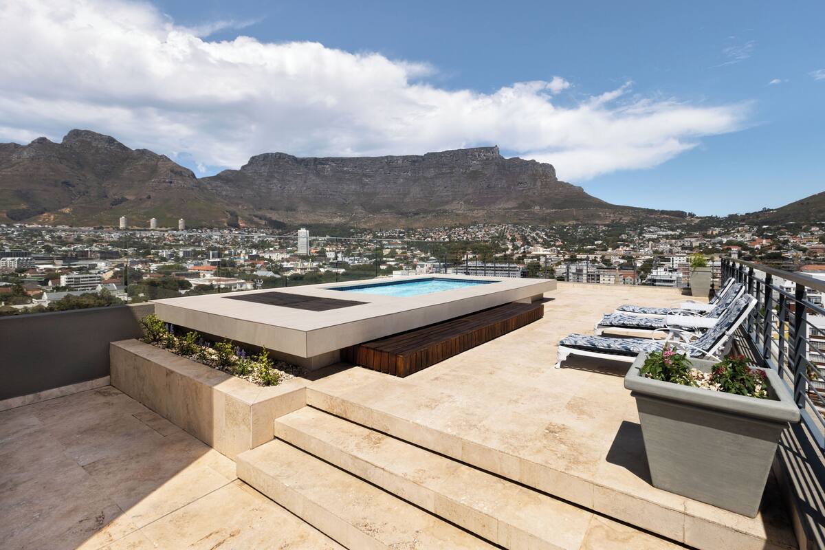 Terraza en la azotea con piscina, escalones que bajan a la piscina, sillas de descanso y vistas a un paisaje urbano y montañas.