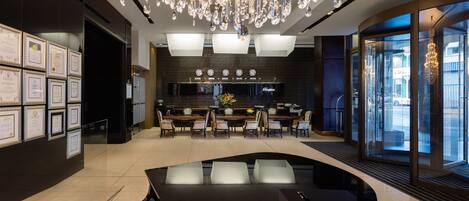 A modern hotel lobby with a large chandelier, a reception desk, framed certificates on the wall, and a seating area with tables and chairs.