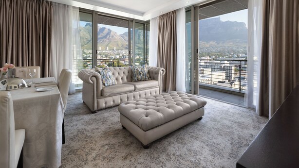 A spacious living room with a large window offering a city view, a tufted sofa, a dining table with a vase of flowers, and a coffee table.