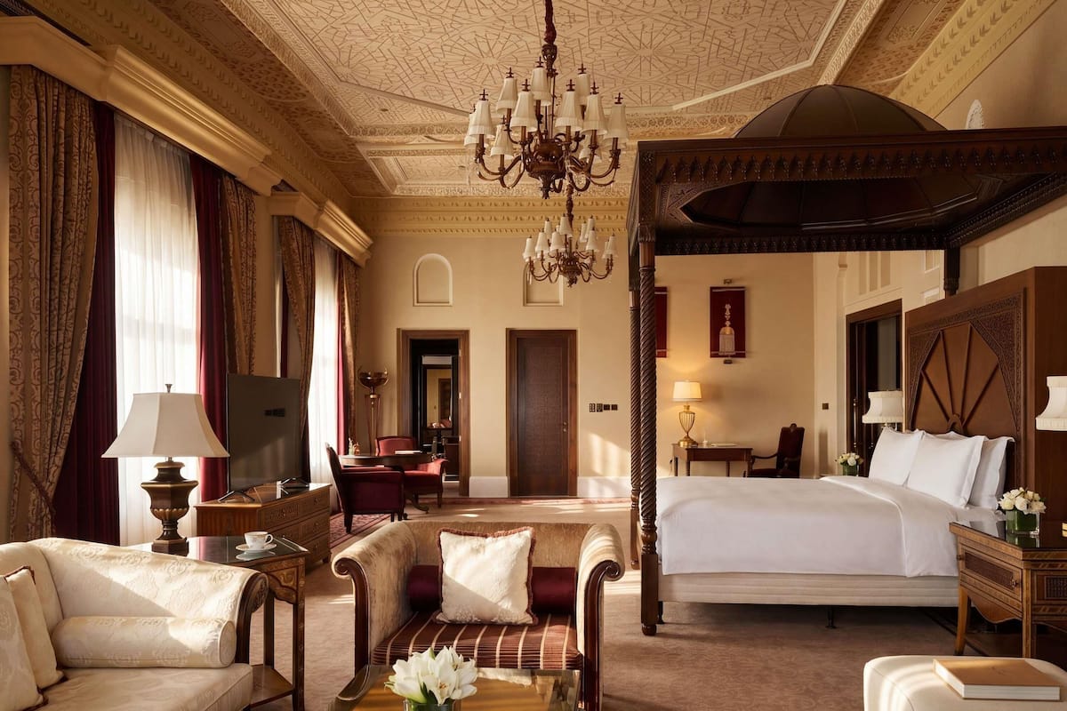 A luxurious hotel room with a four-poster bed, a sitting area with a sofa and armchairs, a dining table, and a chandelier.