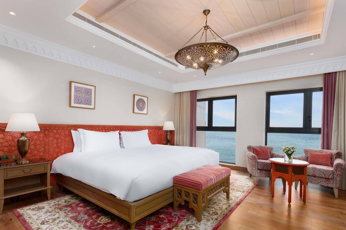 A spacious bedroom with a large bed, a wooden bench, a small table, and a view of the sea through a large window.