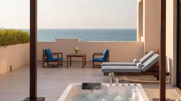 A hot tub with a view of the sea, two lounge chairs, and a table with chairs on a balcony.