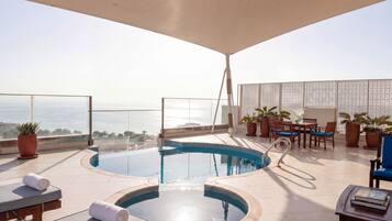 Una piscina infinita en la azotea con sillones de descanso y una mesa, rodeada de plantas en macetas y vistas al océano.