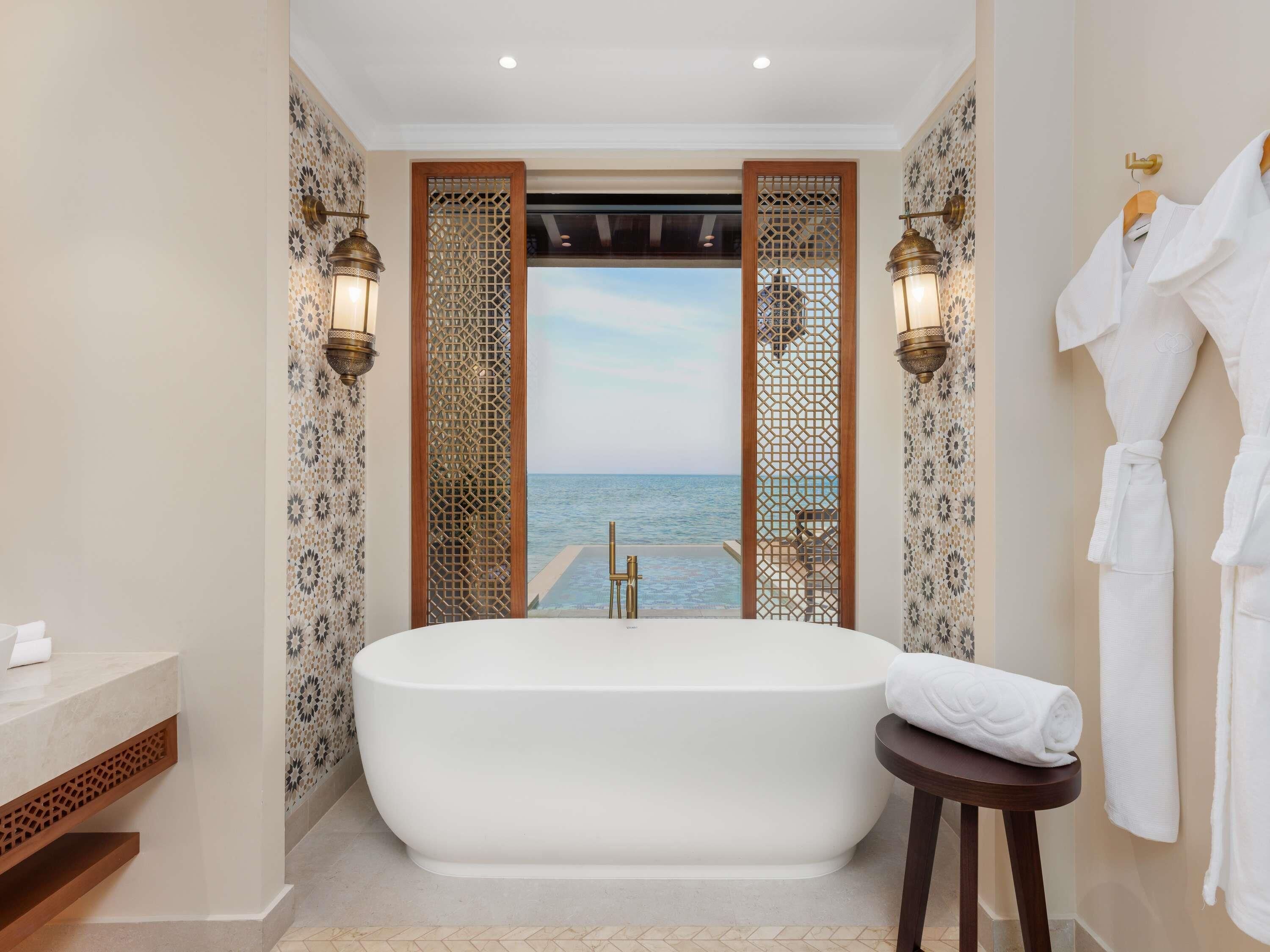 Un baño con una bañera grande, un banco de madera y vistas al mar a través de una celosía decorativa.