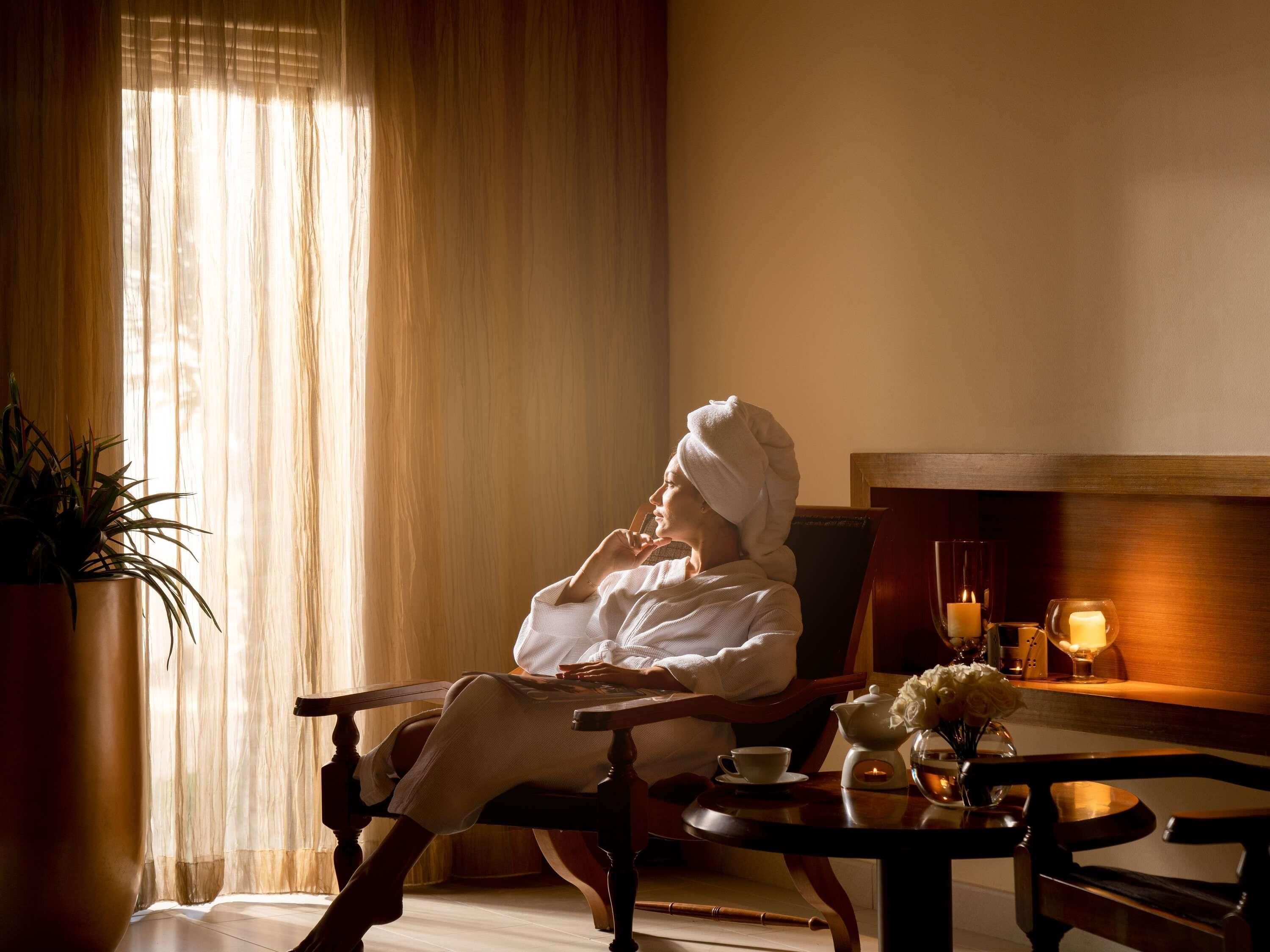 A person seated in a chair with a towel on their head, a table with a teapot, cup, and flowers, and a potted plant.