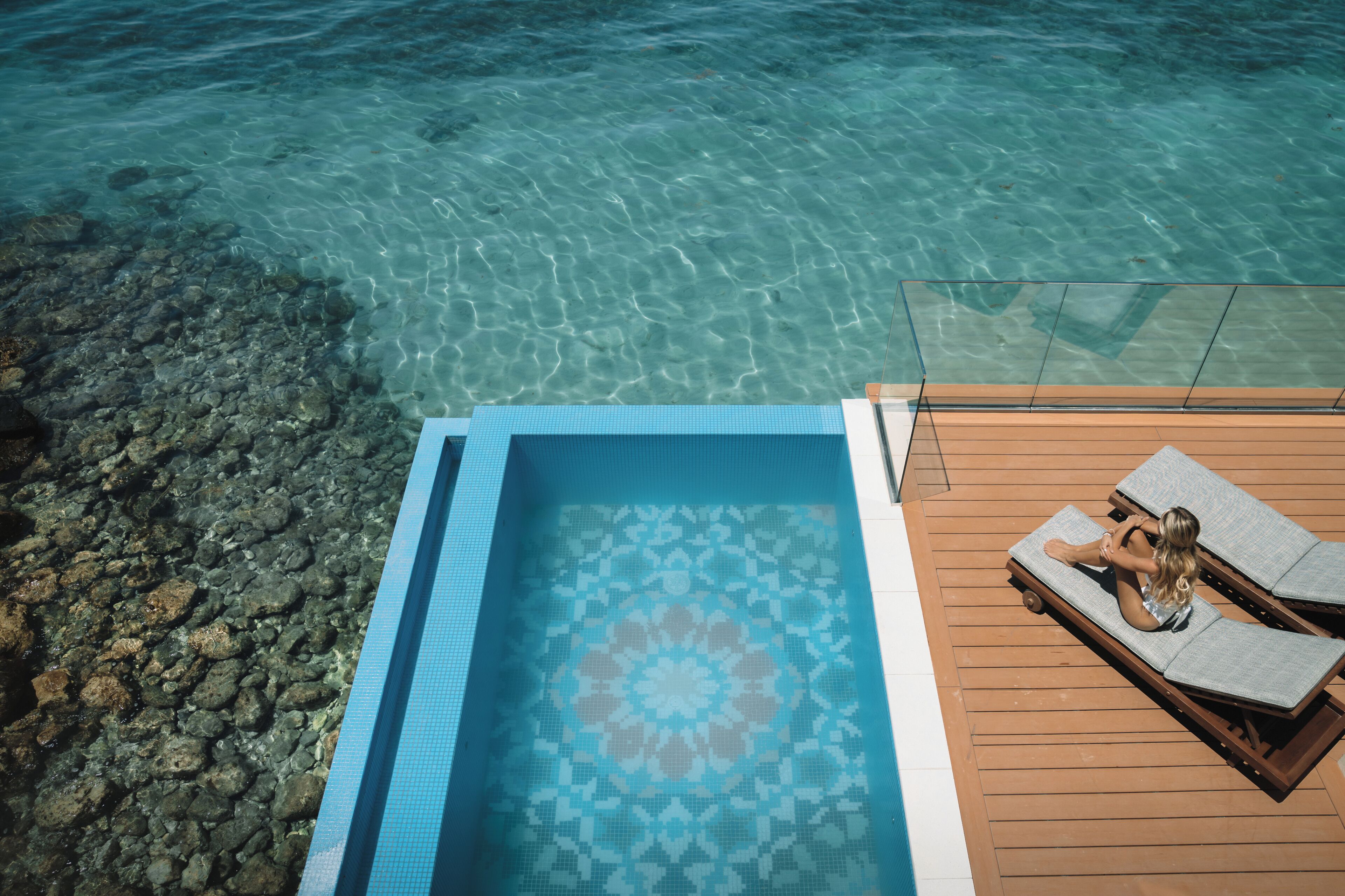 Une piscine aux eaux cristallines, dont le fond est décoré de motifs géométriques en mosaïque, avec une terrasse en bois et des chaises longues.