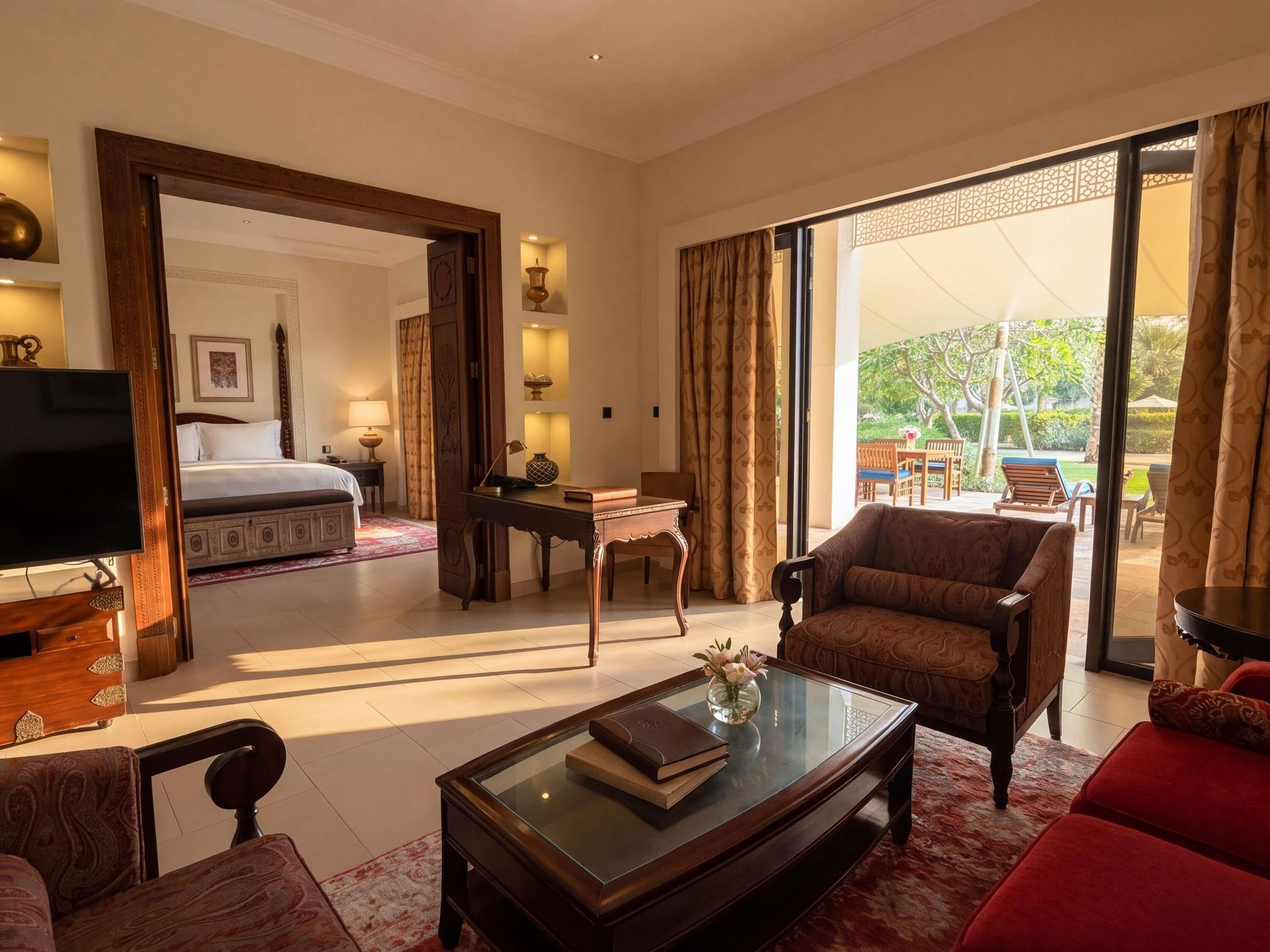 A hotel room with a bed, a TV, a glass coffee table with books, a sofa, and a view of an outdoor area with a patio and chairs.