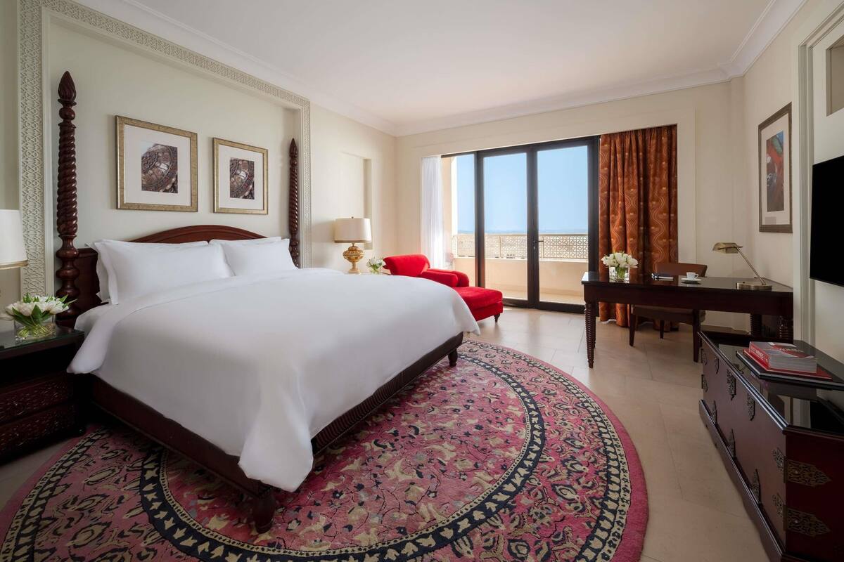 A hotel room with a large bed, a desk, a chair, a TV, and a balcony with a view.