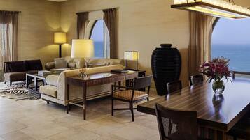 Habitación de hotel con sofá, mesa de comedor, sillas, lámpara, un jarrón grande con flores y vista al mar.