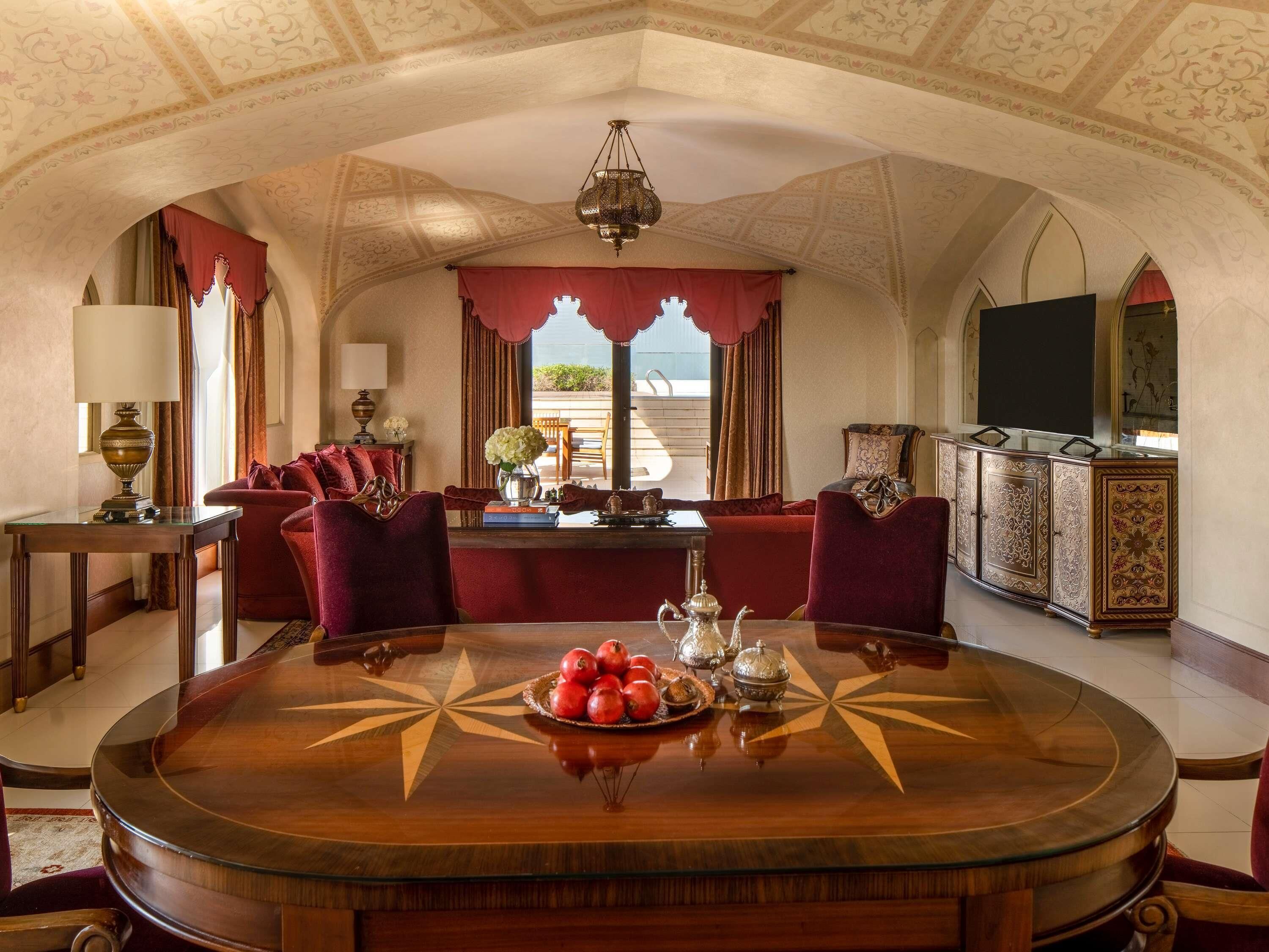 Une chambre luxueuse avec une table ronde en bois, des fauteuils en velours rouge, un lustre et une grande fenêtre avec des rideaux.