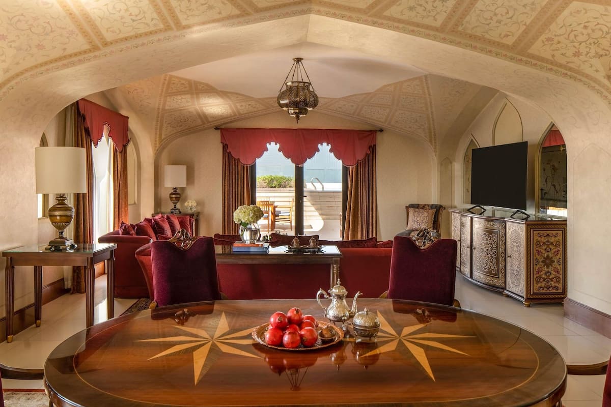 Une chambre luxueuse avec une table ronde en bois, des fauteuils en velours rouge, un lustre et une grande fenêtre avec des rideaux.