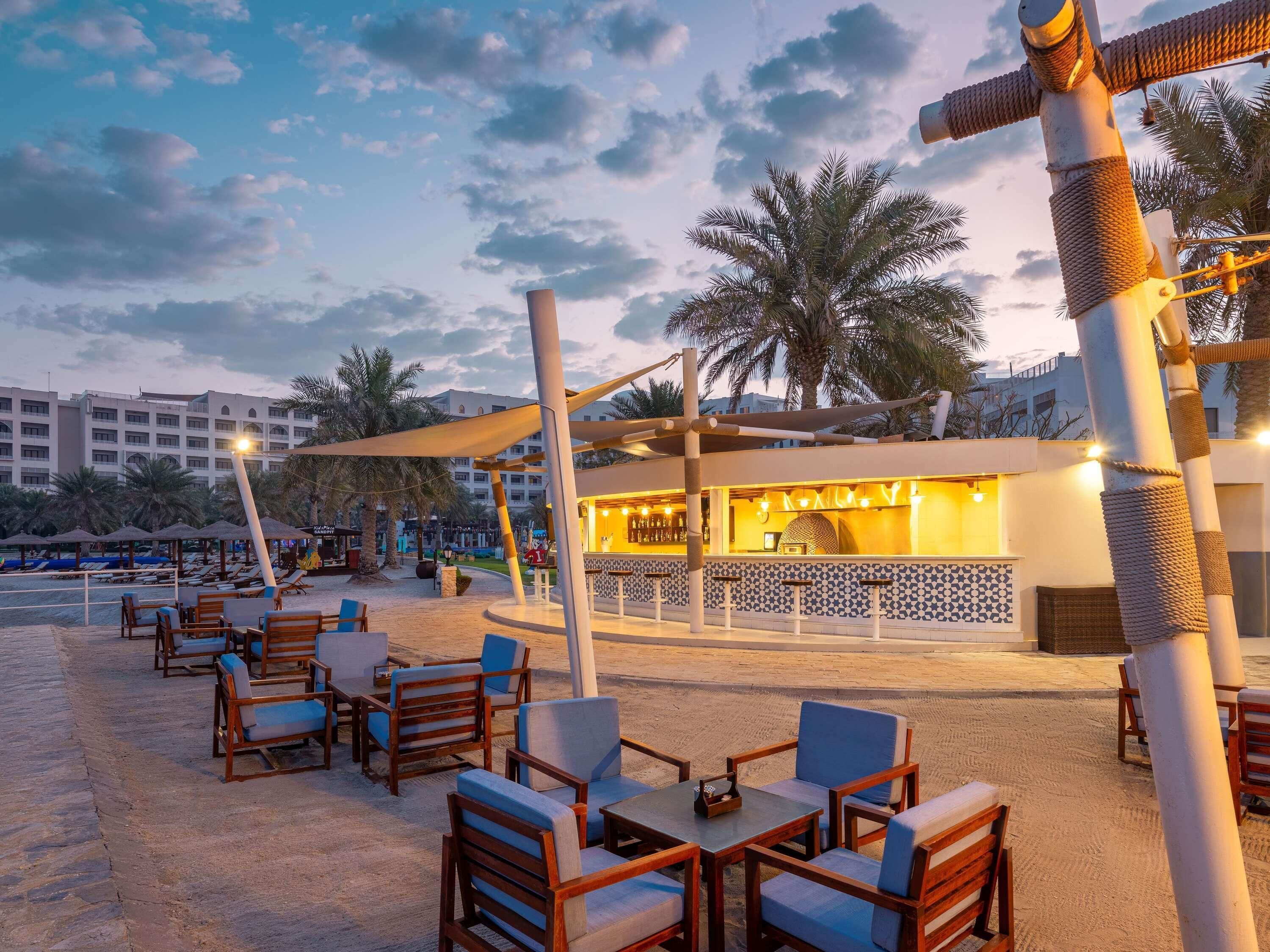 A beachside outdoor seating area with wooden chairs and tables, a bar with a patterned wall, and palm trees in the background.