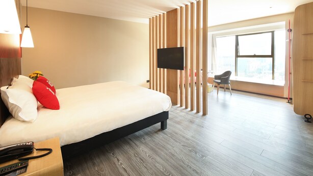 Quarto de hotel moderno com uma cama grande, televisão de tela plana fixada em uma divisória de madeira, piso de madeira e uma ampla janela com vista para a cidade.