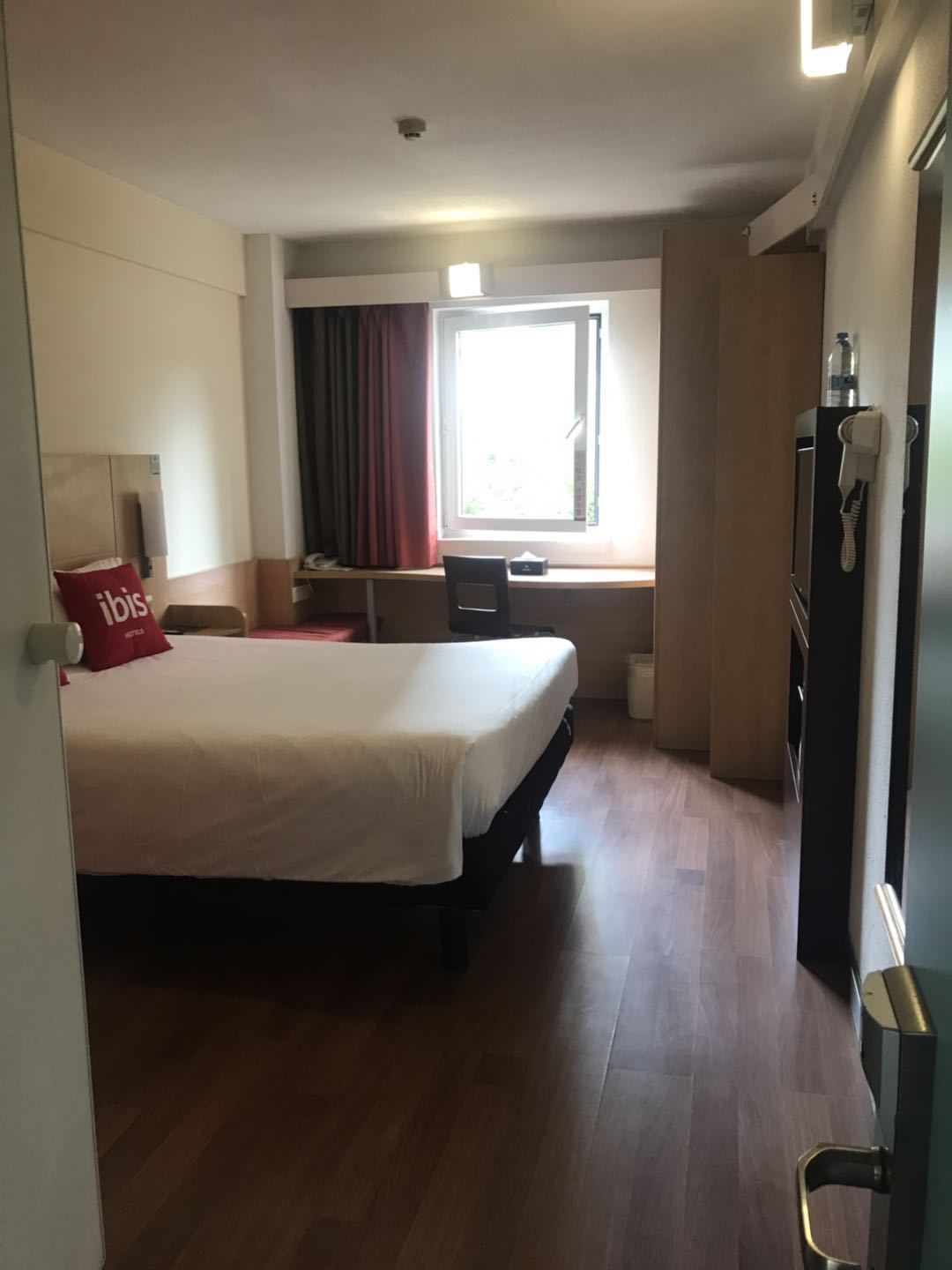 A hotel room with a bed, a desk, a chair, a window with curtains, and a door with a keycard lock.