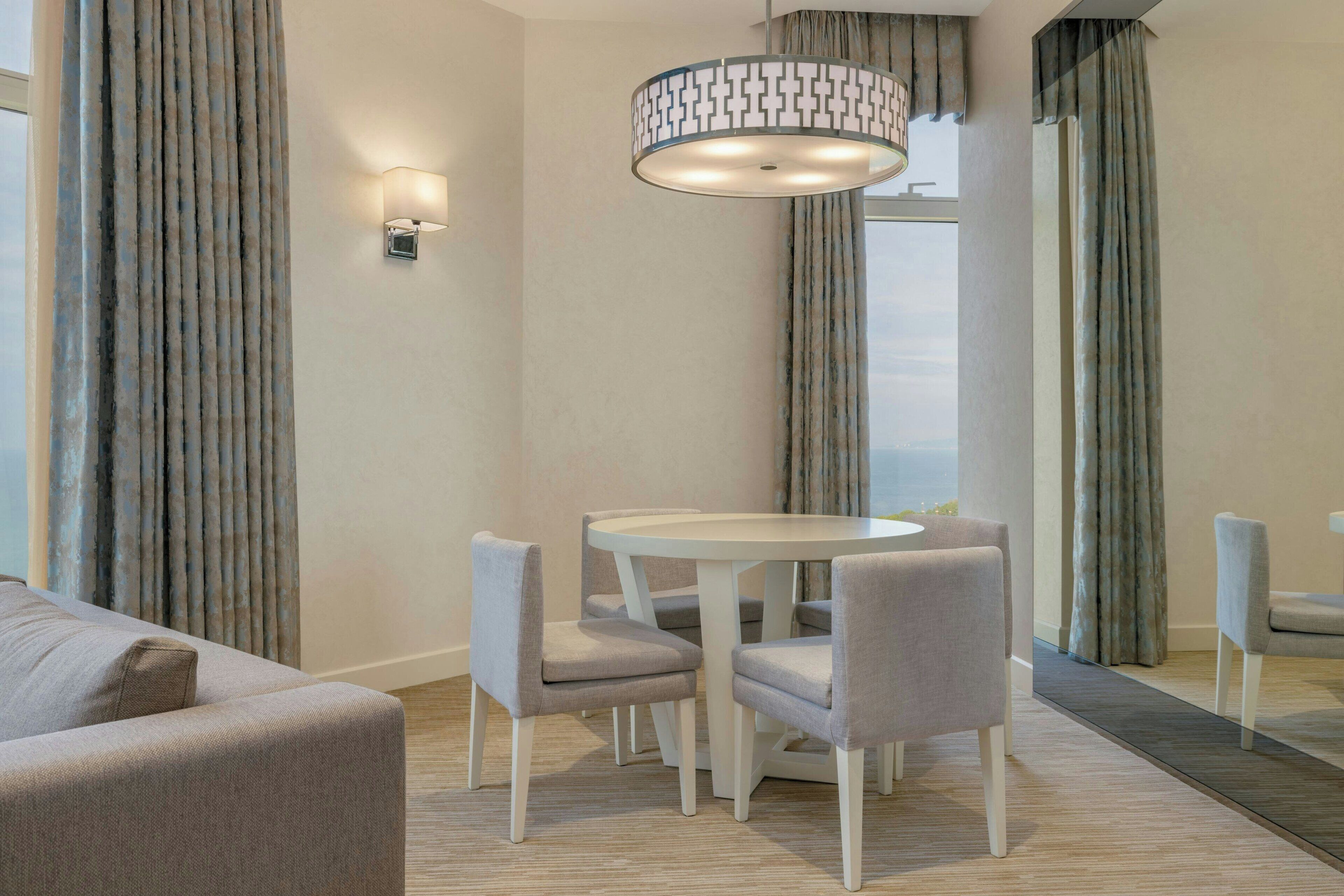 A modern hotel room with a round dining table, four chairs, a sofa, and a large window with curtains.