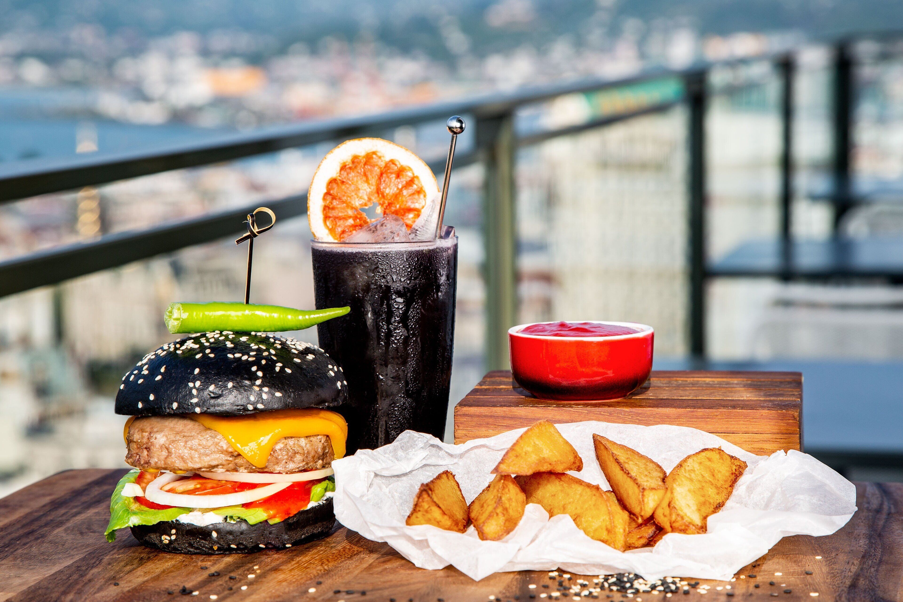 A burger, a drink with a garnish, and fries on a wooden table with a city view in the background.