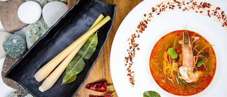 A plate of soup with shrimp and vegetables, accompanied by lime wedges and chili peppers on a wooden table.