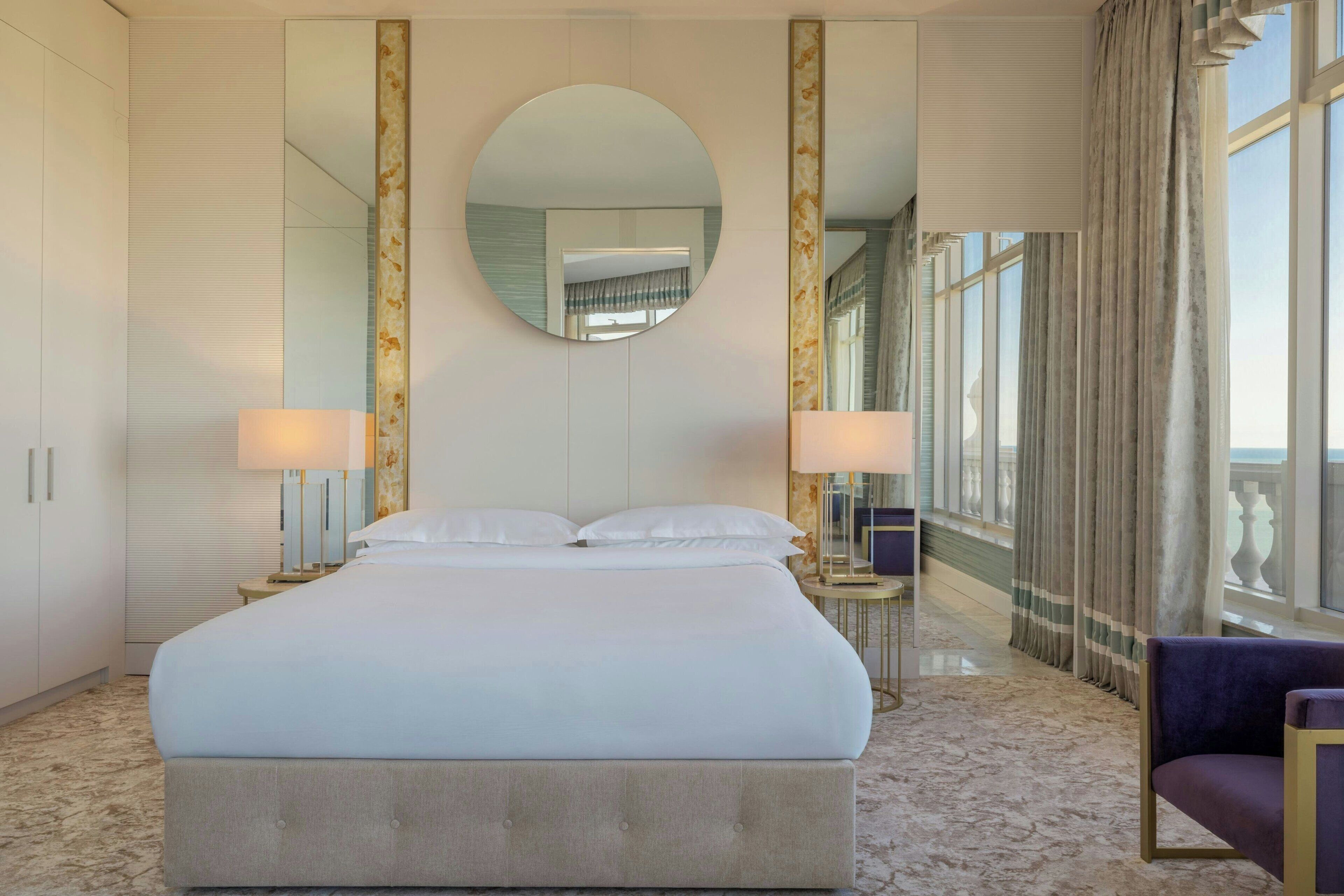 A large bed with a headboard, two bedside lamps, a round mirror, and a view of a balcony through large windows.