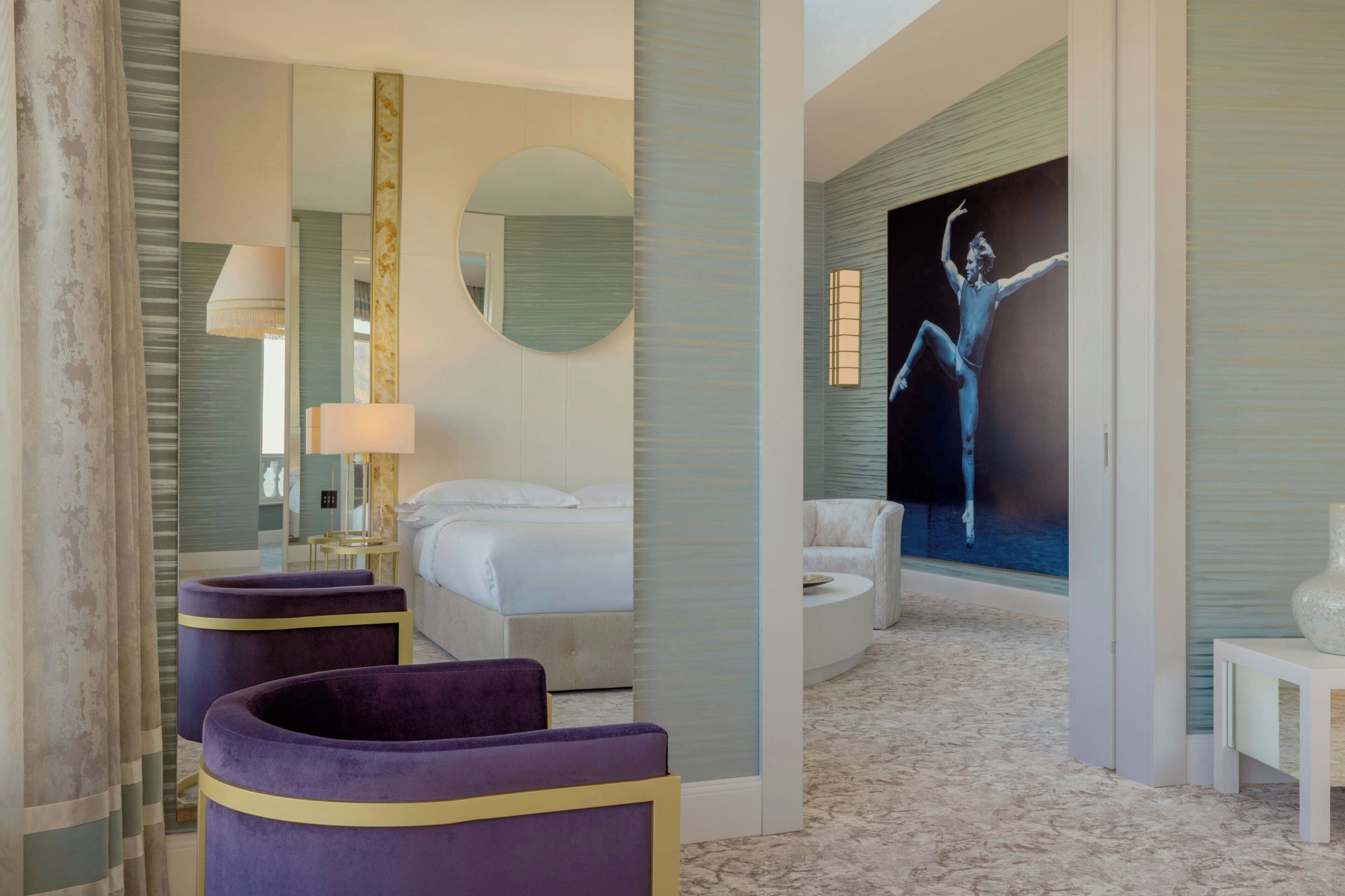 A hotel room with a bed, a round mirror, a wall-mounted lamp, a purple armchair, a white sofa, and a large painting of a dancer.