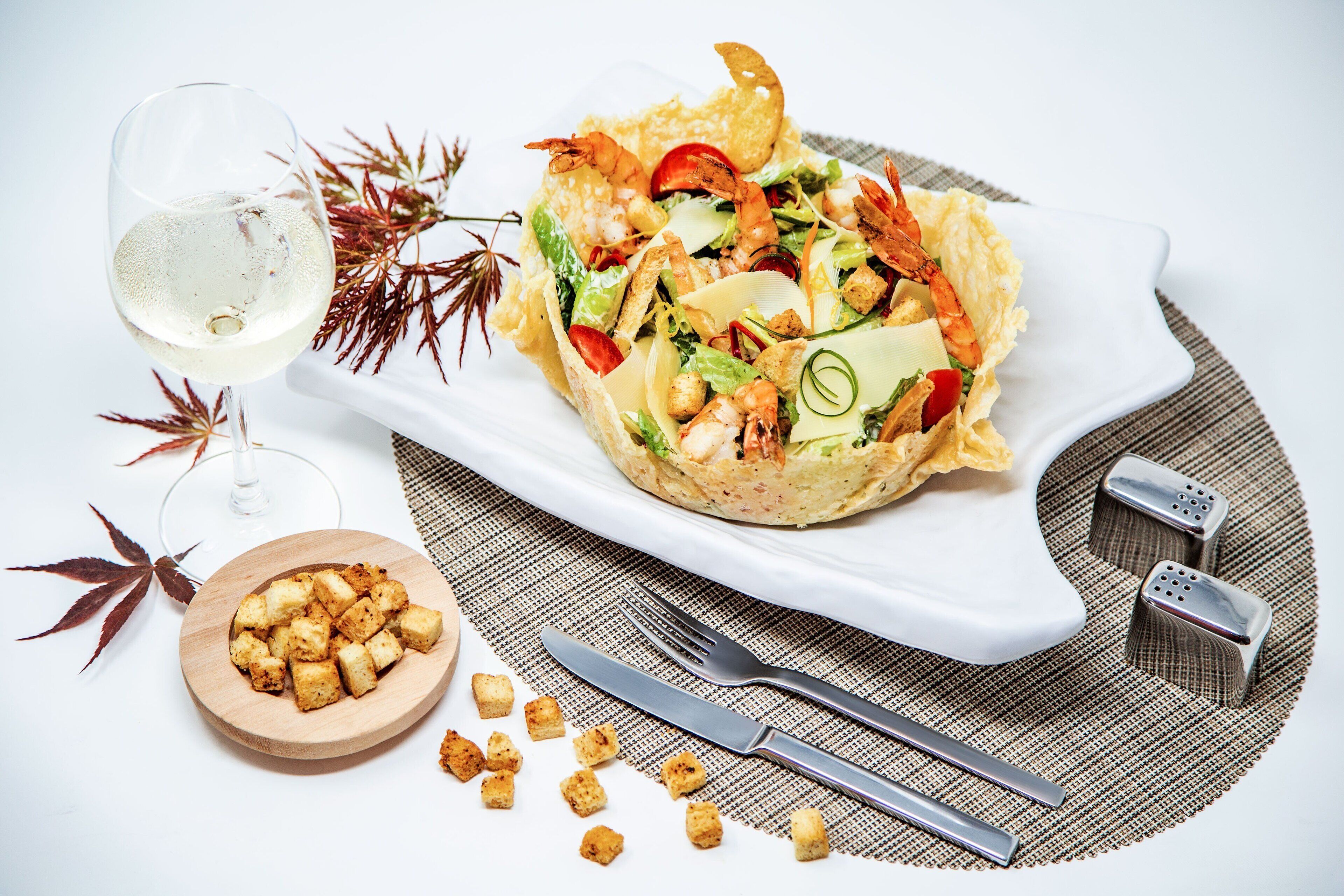A plate of food with a glass of white wine, a wooden bowl of croutons, and a fork and knife on a placemat.