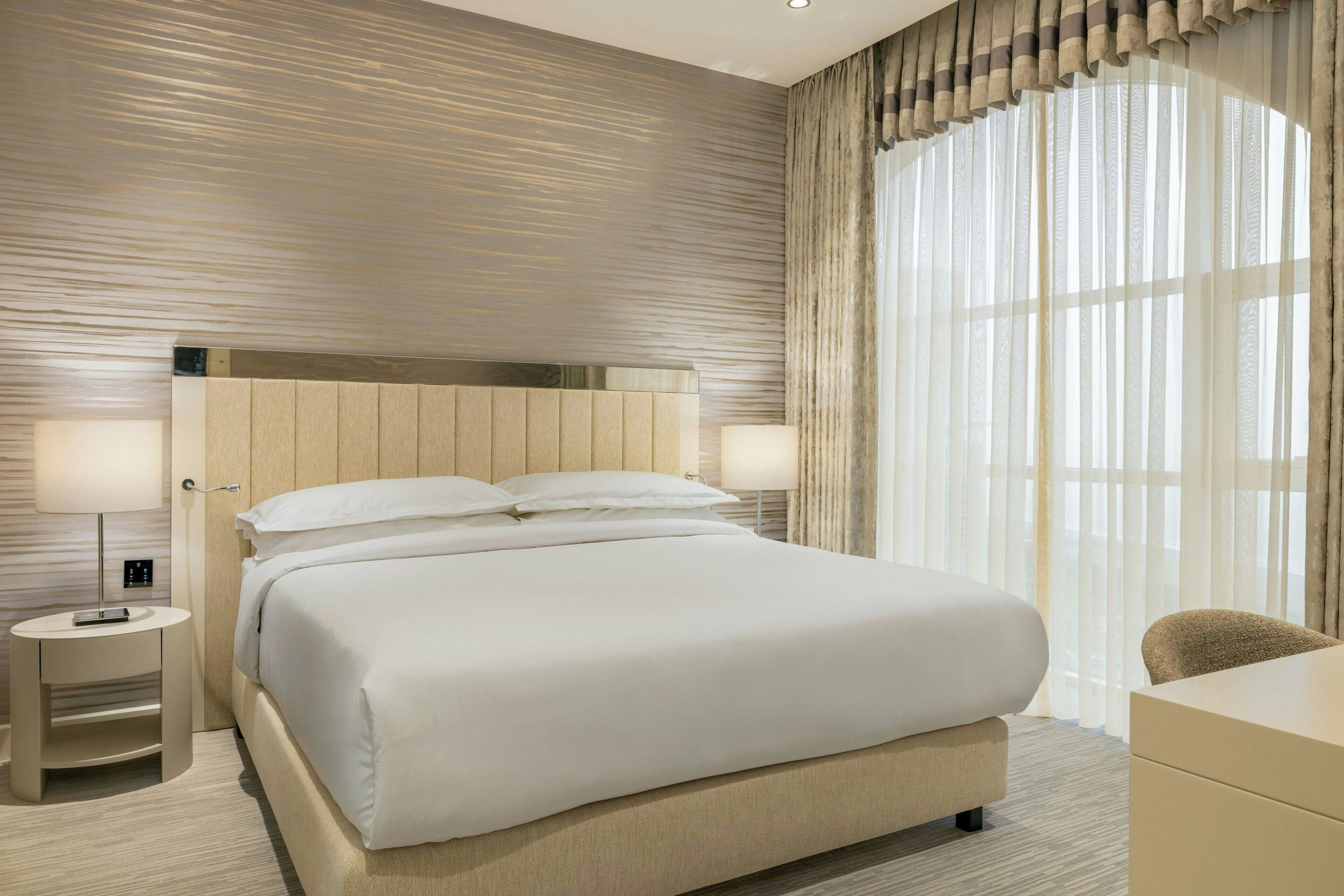 A modern hotel room with a large bed, a desk, and a view of the city through sheer curtains.