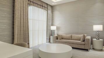 A modern hotel room with a round coffee table, a sofa, two lamps, and a large window with curtains.