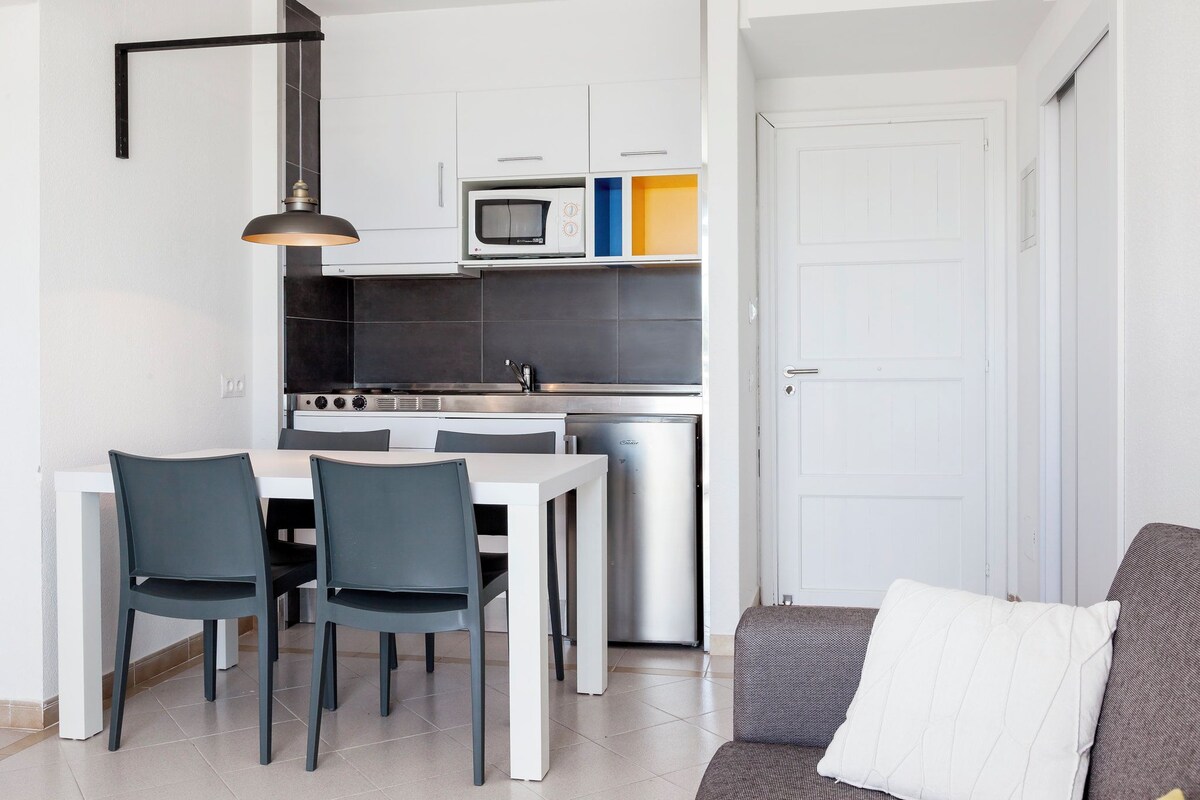 Une cuisine compacte avec des meubles blancs, un four à micro-ondes et un réfrigérateur. On y trouve une table à manger avec des chaises, ainsi qu’un canapé dans l’espace salon.