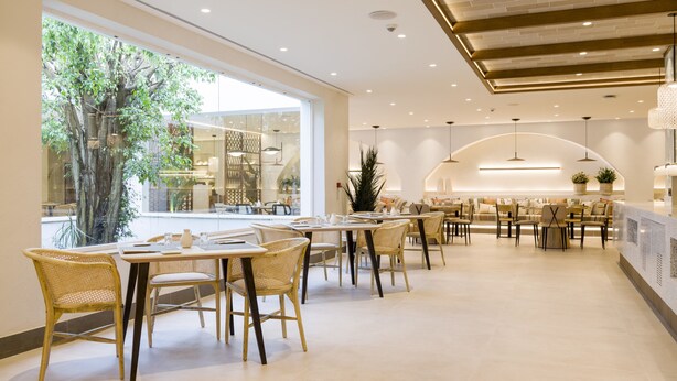 Een moderne eetruimte met rieten stoelen en tafels, grote ramen met uitzicht op bomen, en een centraal zithoek met banken en een hoog plafond met inbouwverlichting.
