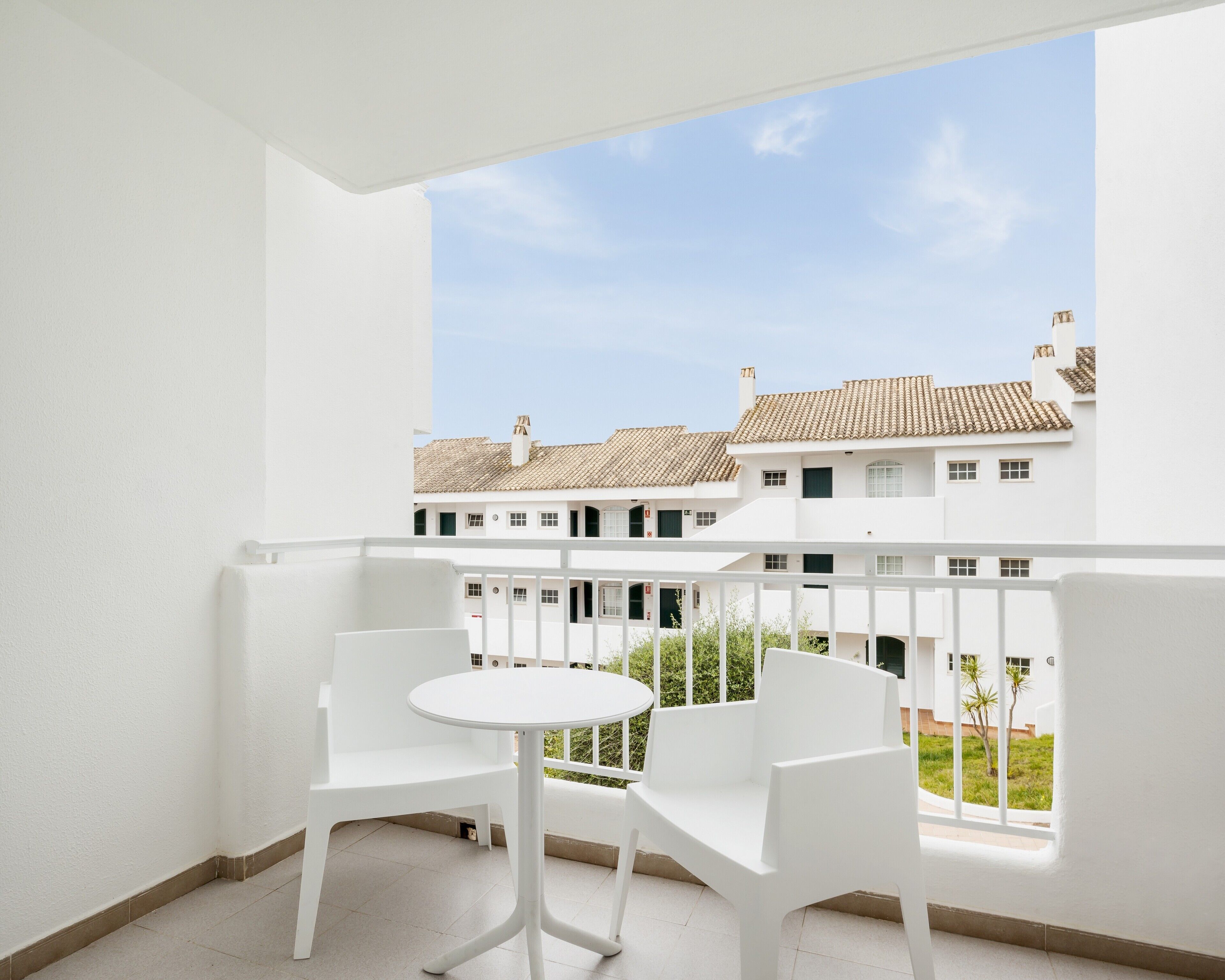 Ein Balkon mit weißen Stühlen und einem kleinen Tisch, mit Blick auf eine Reihung weißer Gebäude mit Ziegeldächern.