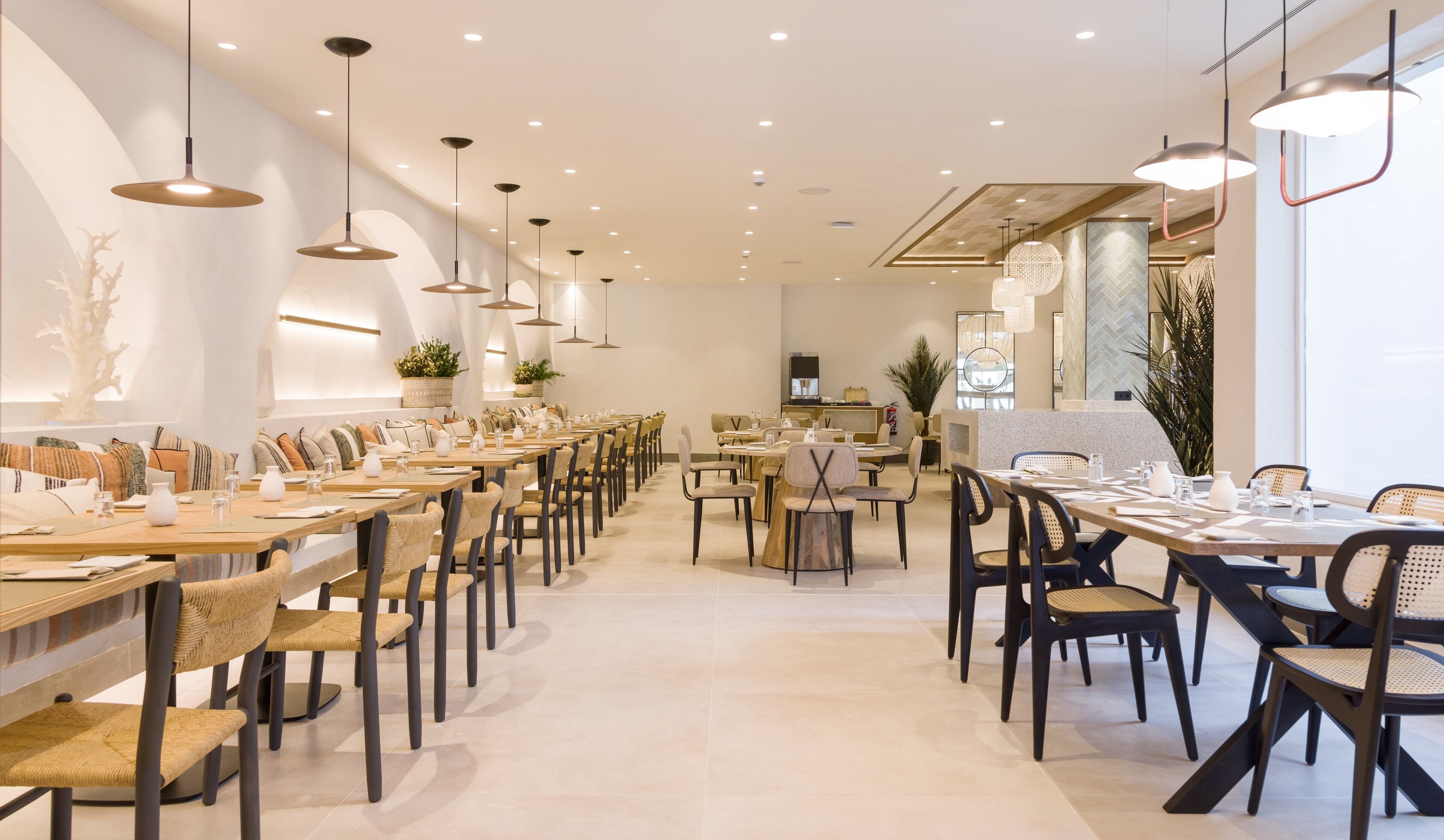 Un vaste espace dédié à la salle à manger, avec plusieurs tables et chaises, des luminaires au plafond et des plantes décoratives.