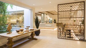 Een moderne hotellobby met een receptiebalie, een eetgedeelte met tafels en stoelen, en een grote kamerplant.