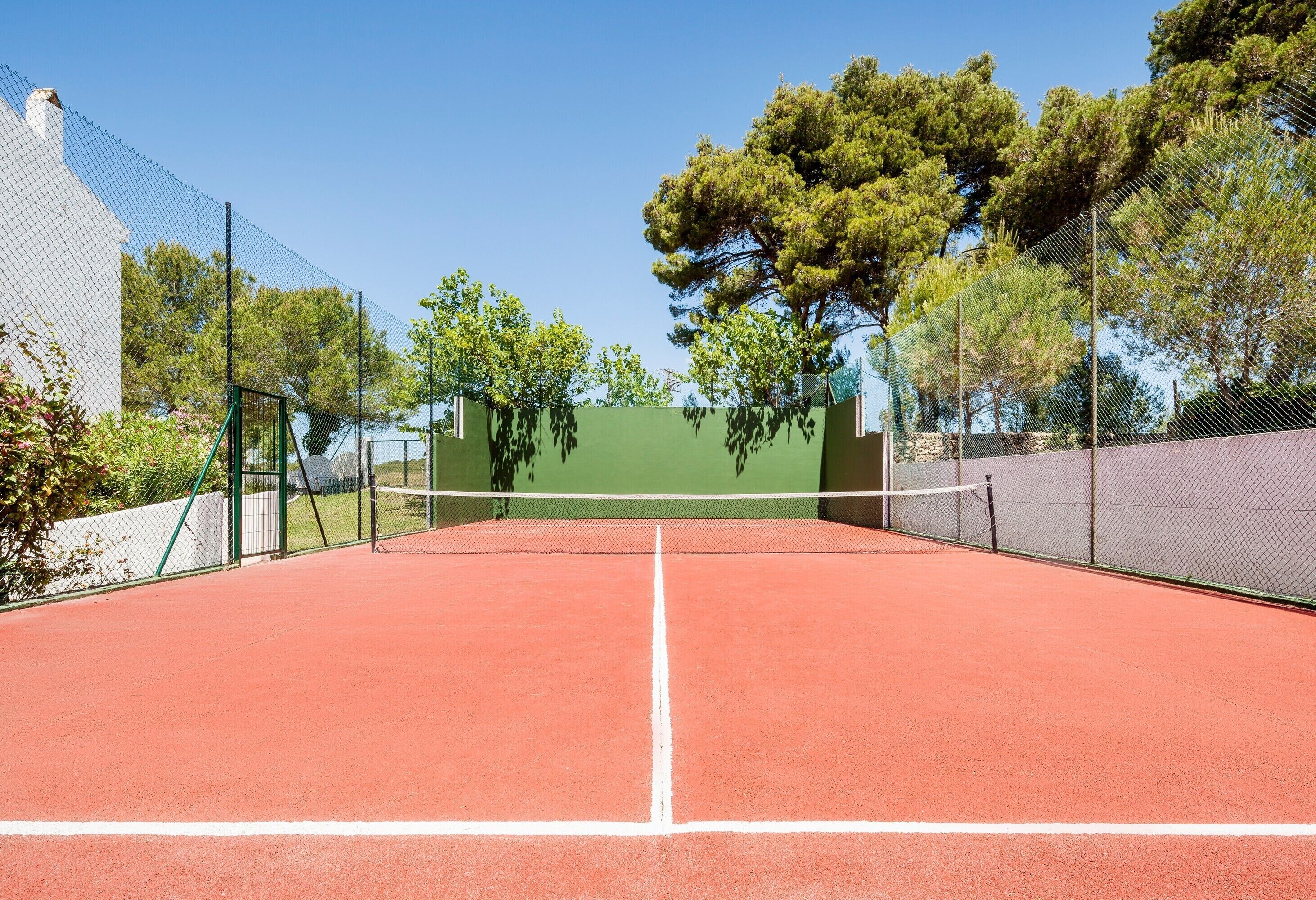 A tennis court with a net, surrounded by green fencing and trees.
