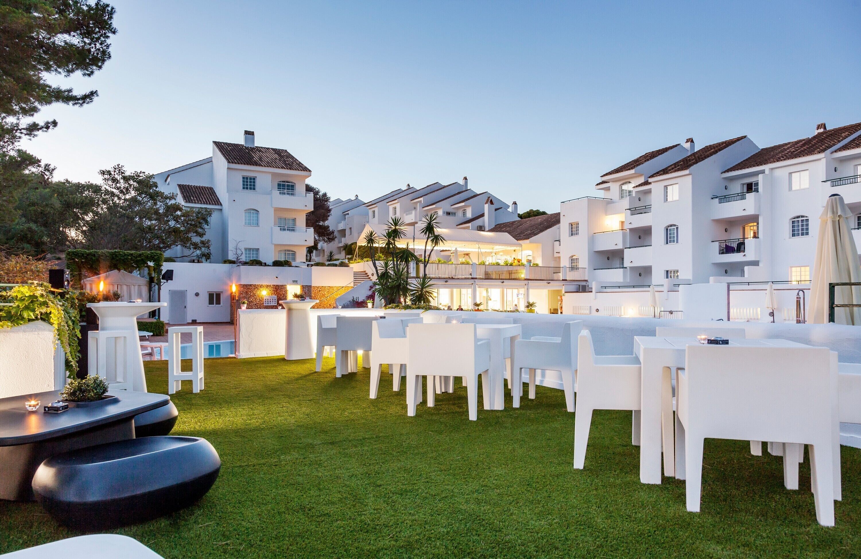 Een verzorgd buitenterras met witte tafels en stoelen, omgeven door weelderig groen gras, grenzend aan een modern gebouw met meerdere balkons.