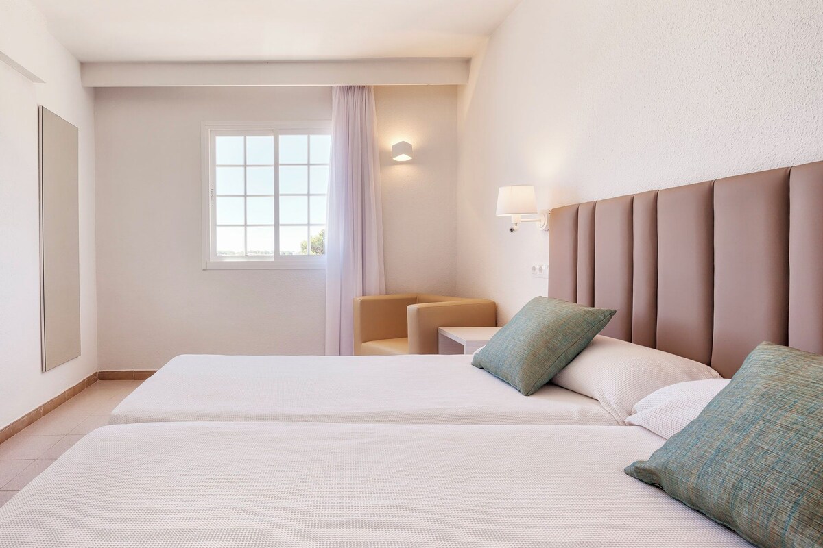 A hotel room with two beds, a window with curtains, a wall-mounted lamp, and a seating area with chairs.