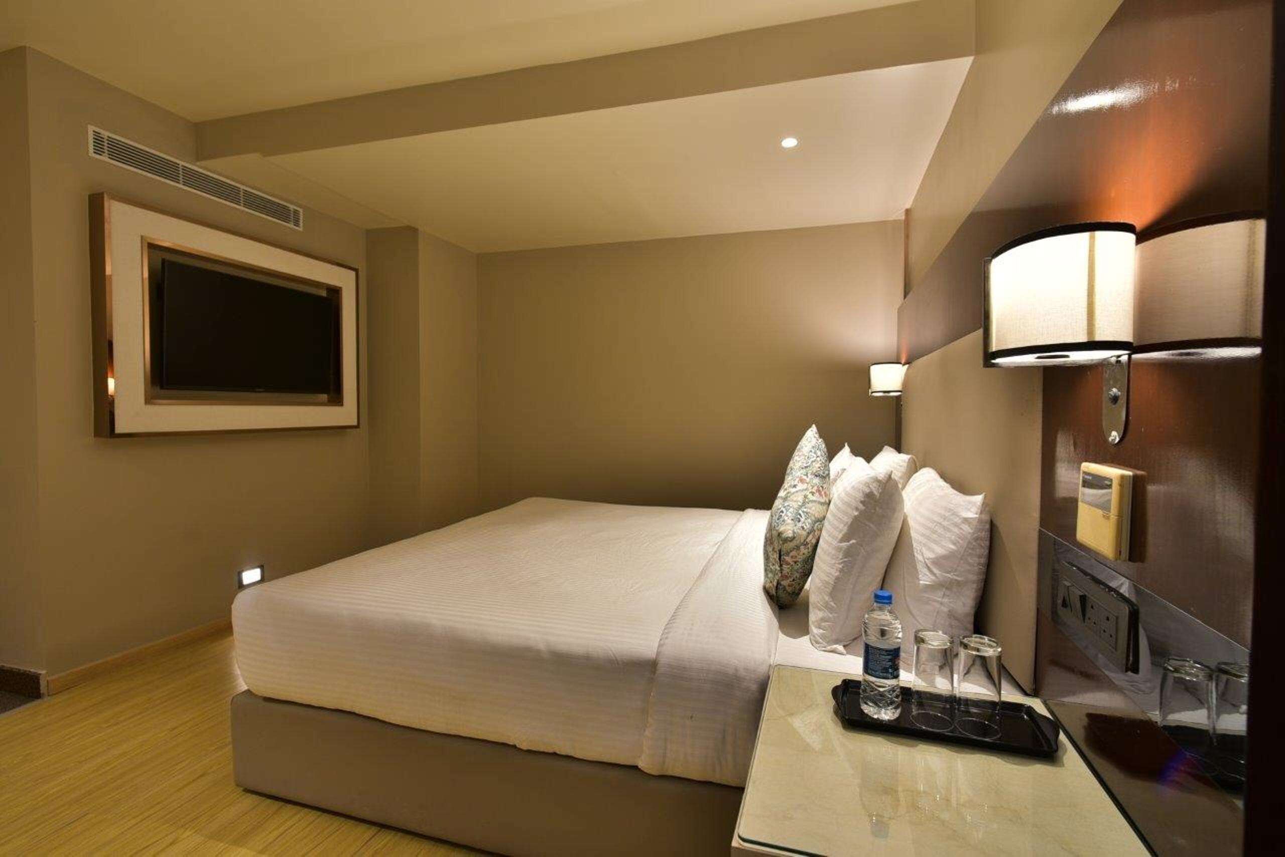 A hotel room with a bed, bedside table with a remote control, water bottles, and a lamp.