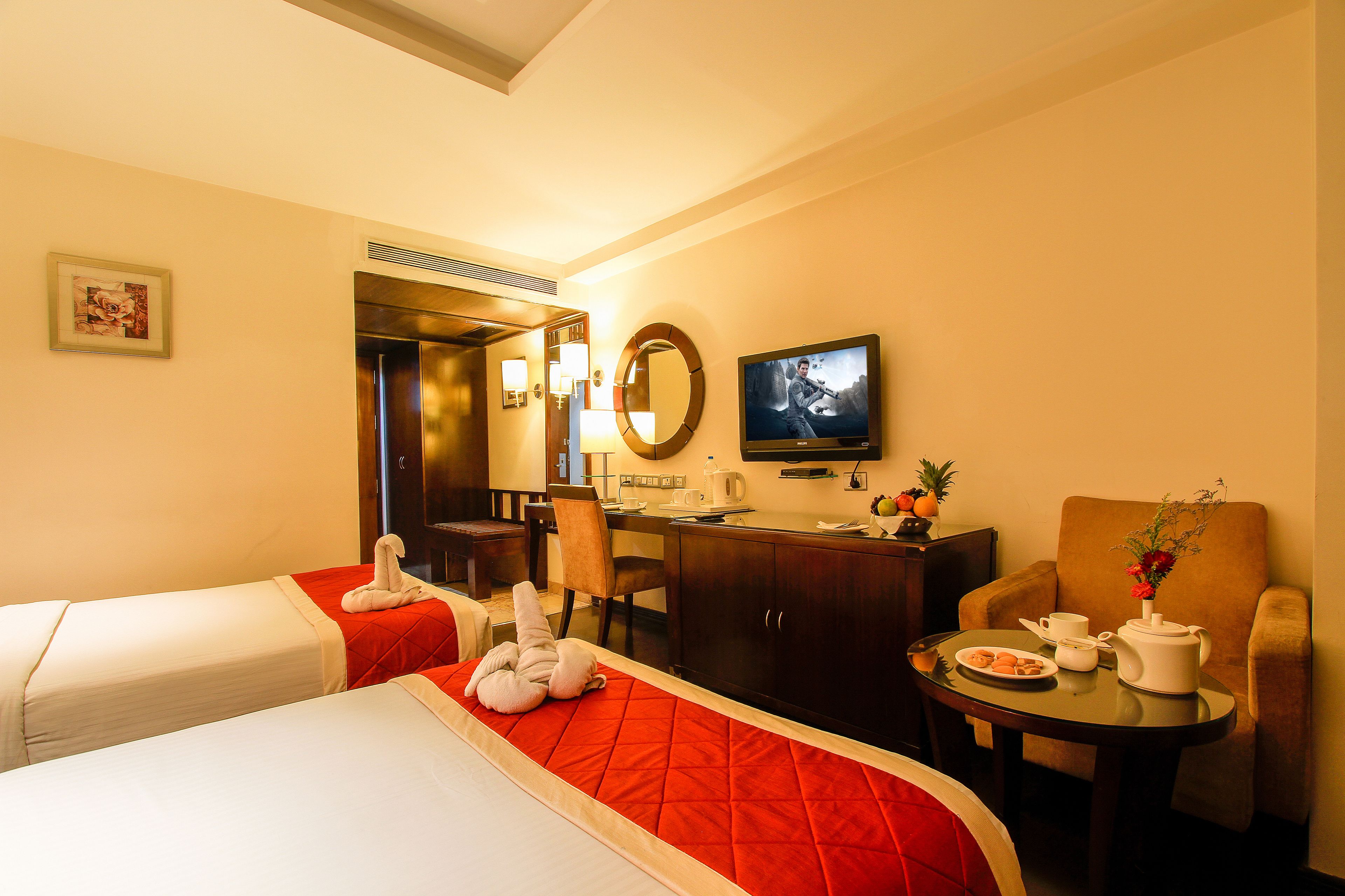 A hotel room with two beds, a desk with a chair, a television, a small table with a vase, and a dining table with a plate of cookies.