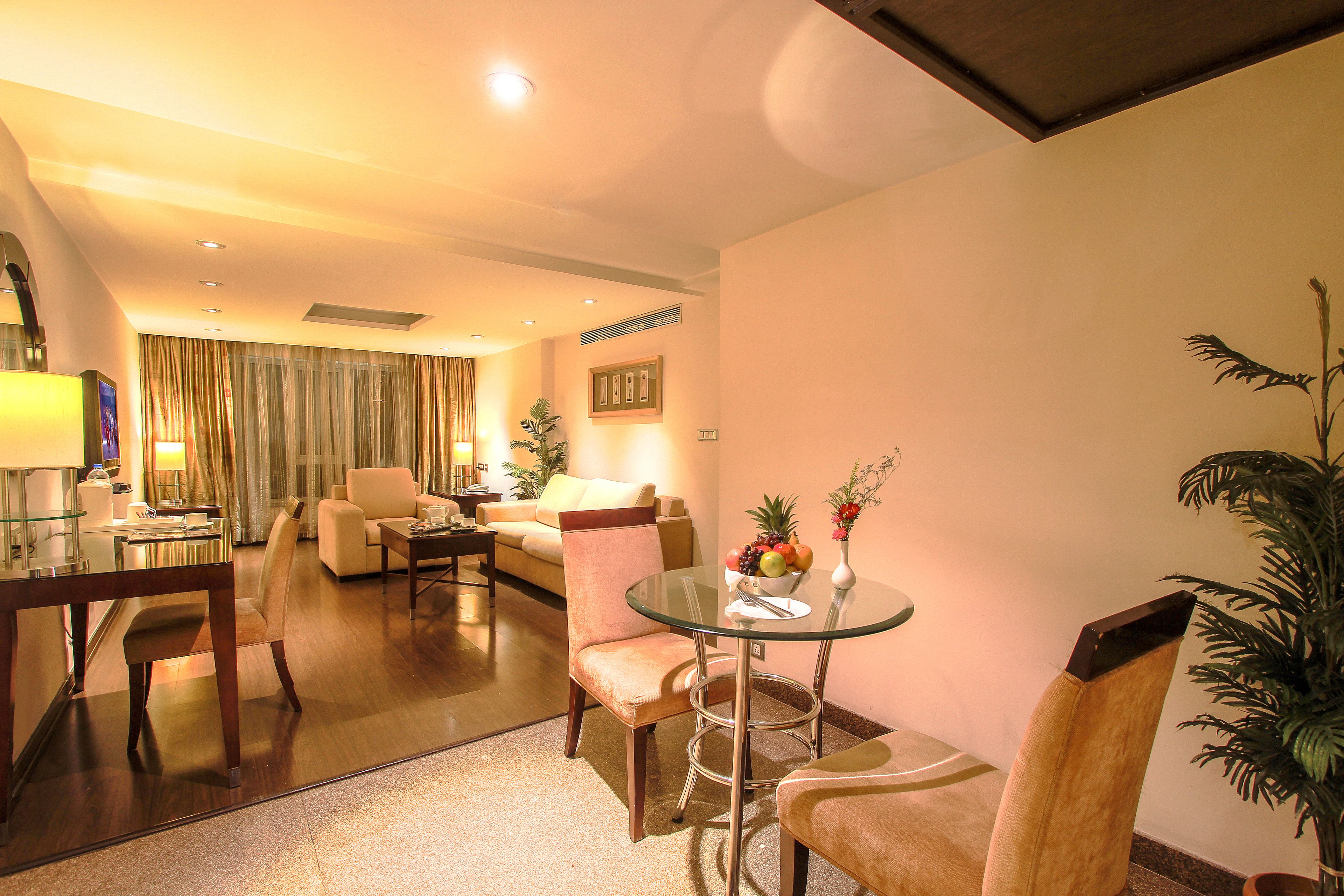 A hotel room with a dining area, a sofa, a glass table with a fruit bowl, and a potted plant.