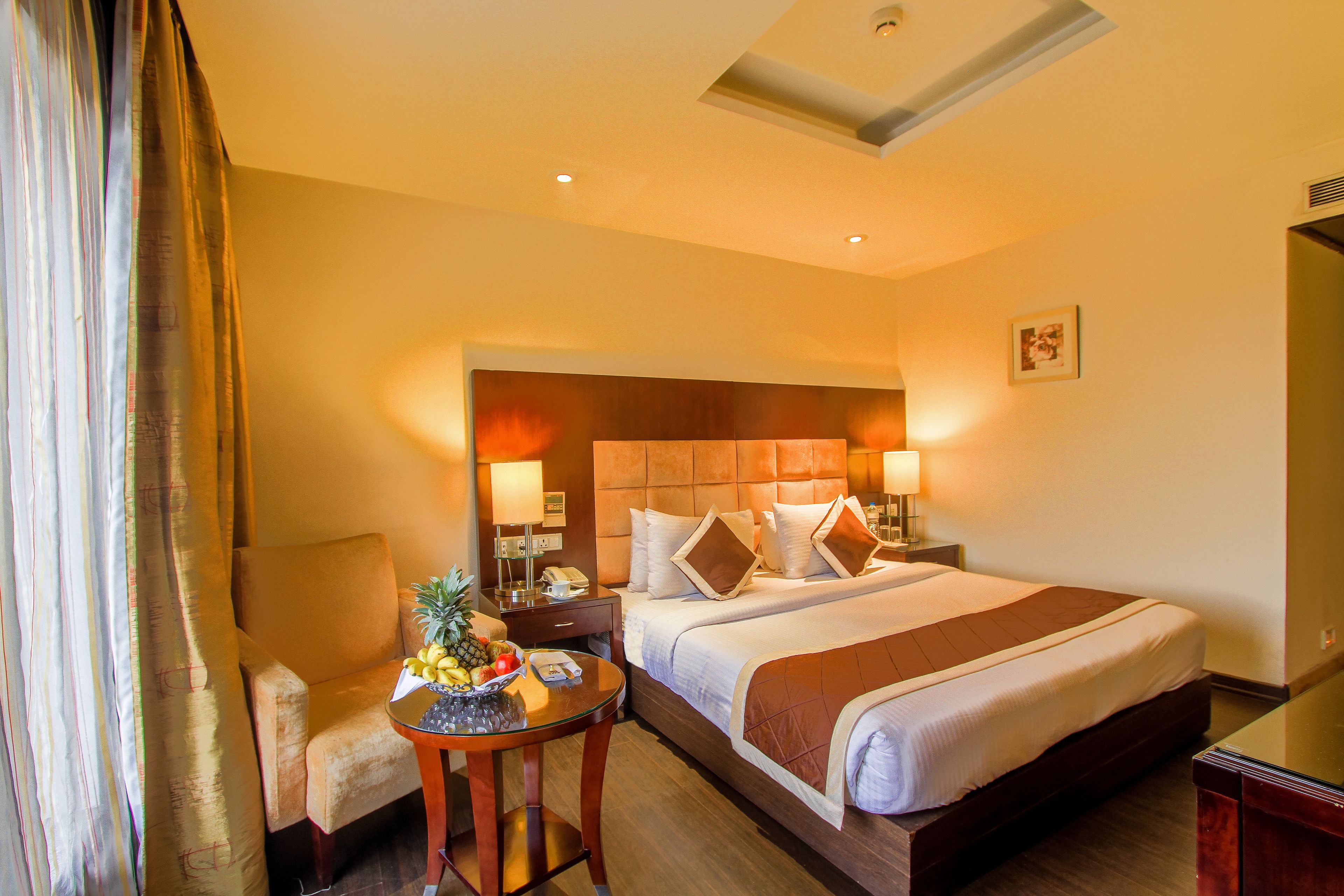 A hotel room with a large bed, a bedside table with a fruit bowl, a chair, and a desk with a lamp.