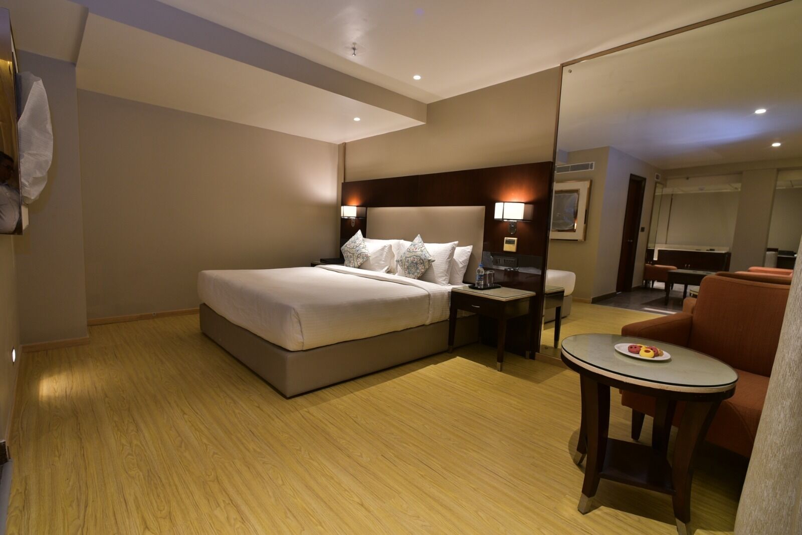 Una habitación de hotel con una cama grande, una mesita de noche con una lámpara, una pequeña mesa redonda con un plato de comida y un área de descanso con una silla.