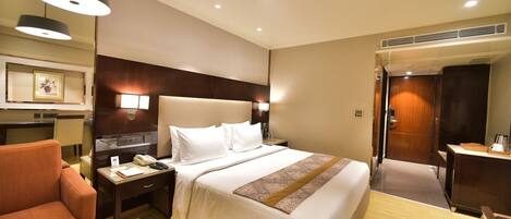 A hotel room with a bed, a desk, a chair, a bedside table, a lamp, and a wall-mounted TV.