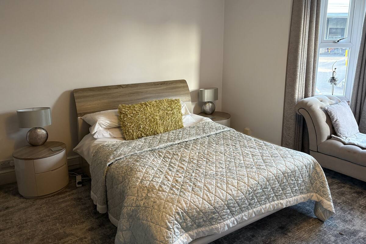 Quarto com cama, mesas de cabeceira, luminária, cadeira e janela com cortinas.