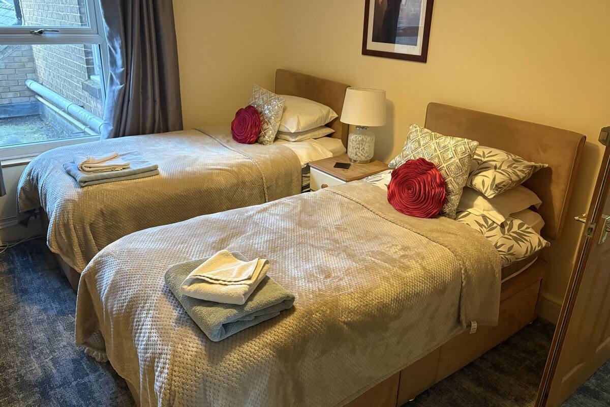Quarto de hotel com duas camas, cada uma com manta e almofadas bege, mesa de cabeceira com abajur e quadro na parede.