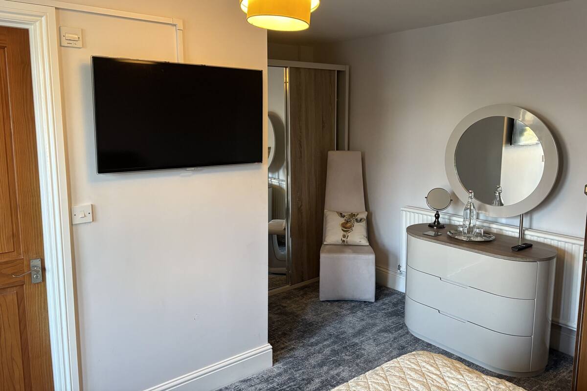 Quarto moderno com televisão de tela plana, espelho redondo, cômoda e um pequeno banquinho.